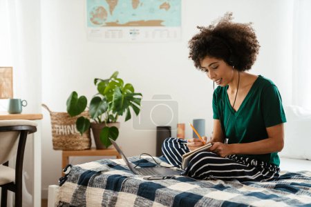 Foto de Mujer joven afroamericana tomando notas mientras está sentada en la cama y usando el portátil en casa - Imagen libre de derechos