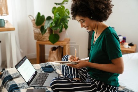 Téléchargez les photos : Africaine américaine jeune femme prendre des notes tout en étant assis dans le lit et en utilisant un ordinateur portable à la maison - en image libre de droit