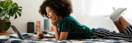 Photo for African american young woman with curly hairstyle lying in bed and using laptop at home - Royalty Free Image