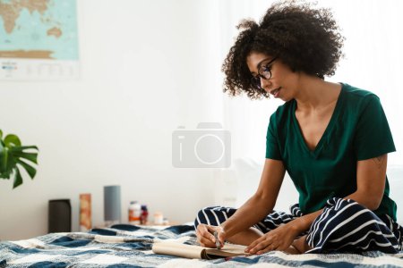 Photo for African american young woman with curly hairstyle sitting in bed and drawing in sketchbook at home - Royalty Free Image