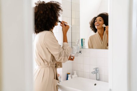 Photo for African american young woman looking in mirror and applying makeup with cosmetic brush at home - Royalty Free Image