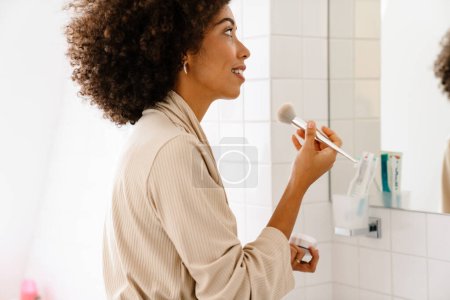 Foto de Mujer joven afroamericana mirando en el espejo y aplicando maquillaje con cepillo cosmético en casa - Imagen libre de derechos