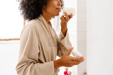 Photo for African american young woman with curly hairstyle smiling and holding face cream in bathroom - Royalty Free Image