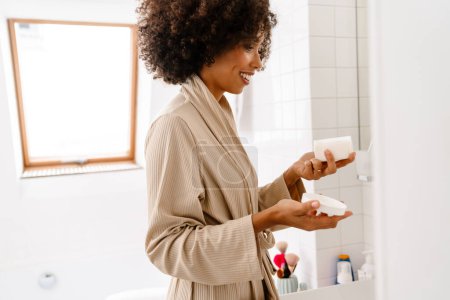 Téléchargez les photos : Afro-américaine jeune femme avec bouclé coiffure souriant et tenant crème visage dans la salle de bain - en image libre de droit