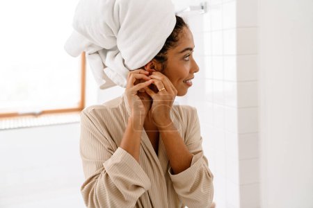 Photo for African american young woman with towel on head smiling and wearing earrings in bathroom - Royalty Free Image