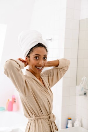 Foto de Mujer joven afroamericana con toalla en la cabeza sonriendo y mirando a la cámara en el baño - Imagen libre de derechos