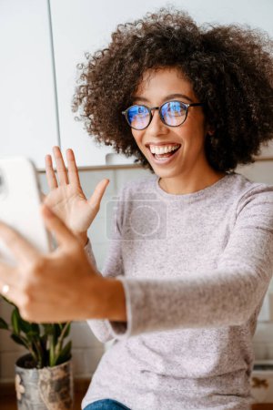 Téléchargez les photos : Africaine américaine jeune femme avec frisée afro coiffure souriante tout en utilisant un téléphone portable à la maison - en image libre de droit