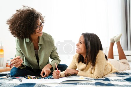 Photo for Black girl and her mother painting in exercise book together on bed at home - Royalty Free Image