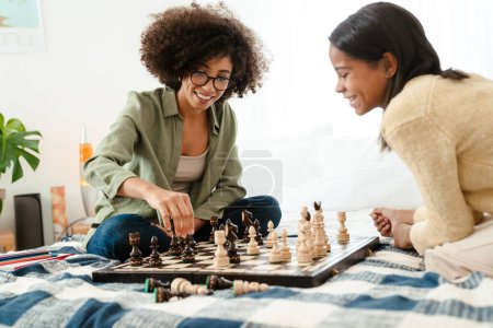 Foto de Mujer afroamericana feliz y su hija adolescente jugando ajedrez mientras está sentada en la cama en casa - Imagen libre de derechos