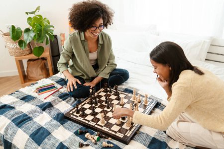 Téléchargez les photos : Heureuse femme afro-américaine et sa fille adolescente jouant aux échecs alors qu'elle était assise au lit à la maison - en image libre de droit