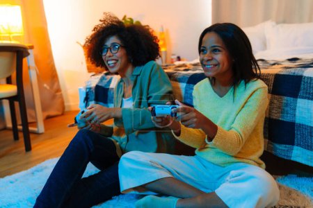 Téléchargez les photos : Heureuse femme afro-américaine et sa fille adolescente jouer à un jeu vidéo avec des contrôleurs à la maison - en image libre de droit