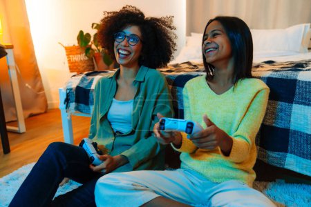 Téléchargez les photos : Heureuse femme afro-américaine et sa fille adolescente jouer à un jeu vidéo avec des contrôleurs à la maison - en image libre de droit