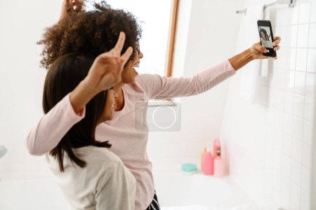 Foto de Chica negra y su madre haciendo gestos mientras toma selfie en el teléfono celular en el baño en casa - Imagen libre de derechos