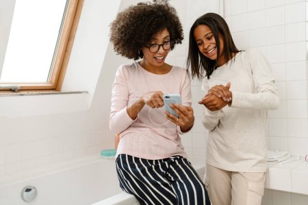 Téléchargez les photos : Heureuse femme afro-américaine et sa fille adolescente utilisant un téléphone portable dans la salle de bain - en image libre de droit