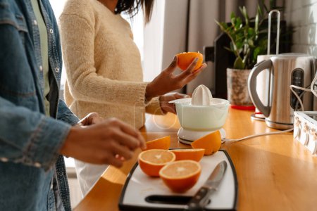 Téléchargez les photos : Heureuse femme afro-américaine et sa fille adolescente faisant du jus d'orange frais dans la cuisine - en image libre de droit