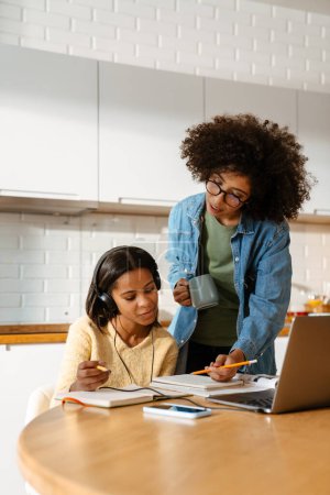 Téléchargez les photos : Jeune femme afro-américaine et sa fille adolescente faisant leurs devoirs ensemble à la maison - en image libre de droit