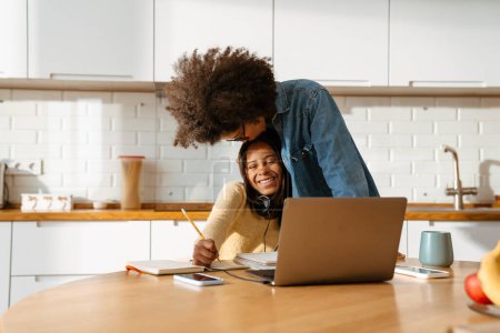 Photo for Young african american woman hugging her teenage daughter while doing homework together at home - Royalty Free Image
