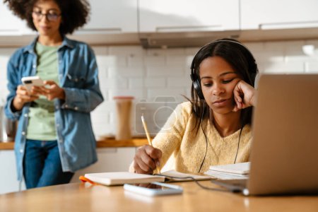Téléchargez les photos : Noir fille écriture dans cahier d'exercices tout en faisant des devoirs avec ordinateur portable à la maison - en image libre de droit