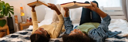 Téléchargez les photos : Fille noire et sa mère riant et lisant des livres tout en étant couché sur le lit à la maison - en image libre de droit