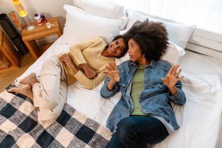 Téléchargez les photos : Fille noire et sa mère riant et parlant tout allongé sur le lit à la maison - en image libre de droit