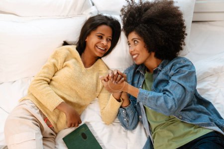 Foto de Chica negra y su madre sonriendo y tomándose de la mano mientras yacía en la cama en casa - Imagen libre de derechos