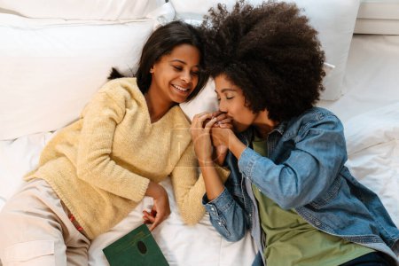Téléchargez les photos : Femme noire embrassant la main de sa fille alors qu'elle était allongée sur le lit à la maison - en image libre de droit