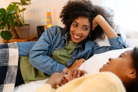 Téléchargez les photos : Fille noire et sa mère riant et parlant tout allongé sur le lit à la maison - en image libre de droit