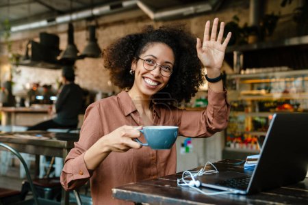 Téléchargez les photos : Jeune femme afro-américaine agitant et buvant du café assis à table dans un café - en image libre de droit