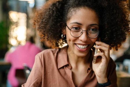 Téléchargez les photos : Jeune femme afro-américaine souriante et parlant sur son téléphone portable dans un café - en image libre de droit