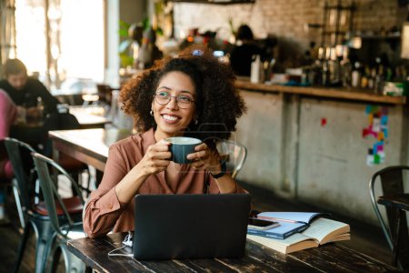 Téléchargez les photos : Jeune femme afro-américaine utilisant un ordinateur portable et boire du café tout en étant assis à table dans un café - en image libre de droit