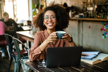 Photo for Young african american woman using laptop and drinking coffee while sitting by table in cafe - Royalty Free Image