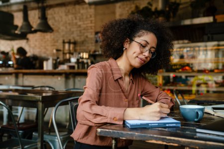 Téléchargez les photos : Jeune femme noire avec afro coiffure prendre des notes dans le livre du planificateur dans le café à l'intérieur - en image libre de droit