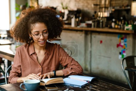 Foto de Joven mujer negra con peinado afro haciendo notas en libro de planificación en la cafetería interior - Imagen libre de derechos