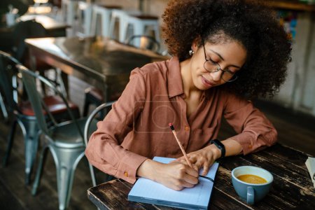 Téléchargez les photos : Jeune femme noire avec afro coiffure prendre des notes dans le livre du planificateur dans le café à l'intérieur - en image libre de droit