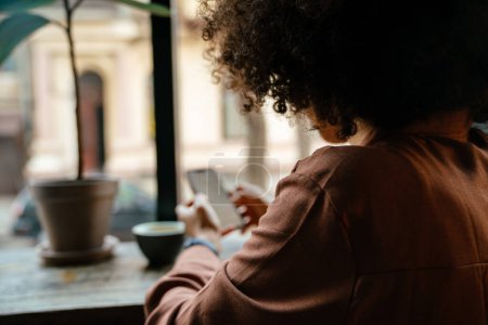 Foto de Joven mujer negra con peinado afro usando teléfono celular y tomando café en la cafetería interior - Imagen libre de derechos