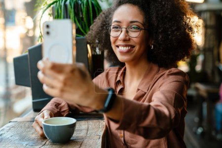 Foto de Joven mujer negra con peinado afro usando teléfono celular y auriculares en la cafetería en interiores - Imagen libre de derechos