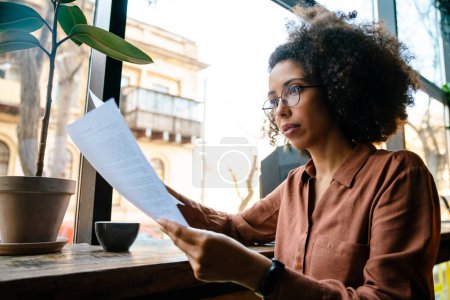 Foto de Joven mujer negra con peinado afro sosteniendo documentos de papel mientras descansa en el café en el interior - Imagen libre de derechos