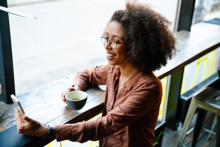 Téléchargez les photos : Jeune femme noire avec une coiffure afro en utilisant un téléphone portable et des écouteurs dans un café à l'intérieur - en image libre de droit