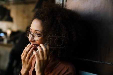 Téléchargez les photos : Jeune femme afro-américaine souriant tout en parlant sur un téléphone portable dans un café - en image libre de droit