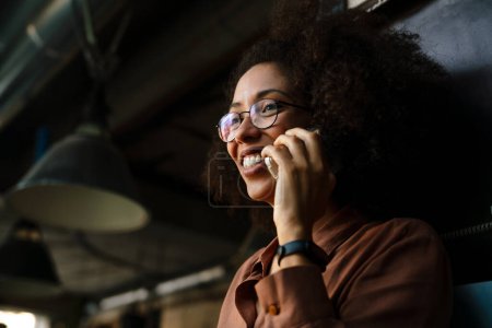 Foto de Joven mujer afroamericana sonriendo mientras habla por teléfono celular en la cafetería - Imagen libre de derechos