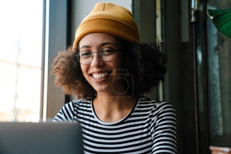 Foto de Joven mujer afroamericana sonriendo y utilizando el ordenador portátil mientras está sentado junto a la mesa en la cafetería - Imagen libre de derechos