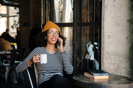 Téléchargez les photos : Jeune femme afro-américaine buvant du thé et utilisant un téléphone portable dans un café - en image libre de droit