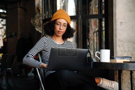 Téléchargez les photos : Jeune femme afro-américaine souriant et utilisant un ordinateur portable assis à table dans un café - en image libre de droit
