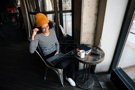 Téléchargez les photos : Jeune femme afro-américaine souriant et utilisant un ordinateur portable assis à table dans un café - en image libre de droit