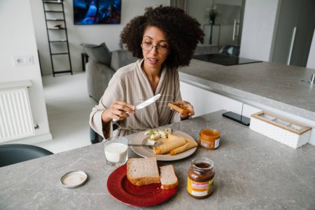 Foto de Mujer negra usando anteojos desayunando mientras está sentada en la mesa en casa - Imagen libre de derechos