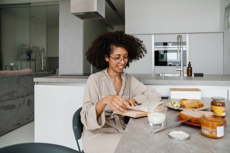 Foto de Mujer negra usando anteojos leyendo libro mientras desayuna en casa - Imagen libre de derechos