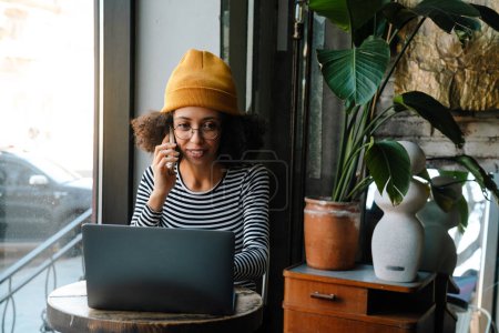 Téléchargez les photos : Jeune femme afro-américaine parlant sur son téléphone portable et utilisant un ordinateur portable assis à table dans un café - en image libre de droit