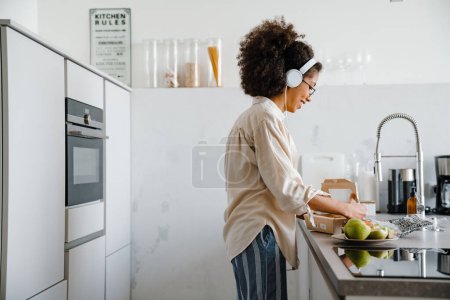 Téléchargez les photos : Jeune femme afro-américaine écoutant de la musique avec des écouteurs tout en faisant le petit déjeuner - en image libre de droit