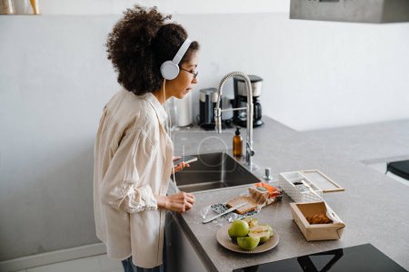 Foto de Mujer joven afroamericana escuchando música con auriculares mientras hace el desayuno - Imagen libre de derechos