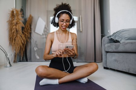 Photo for African american young woman listening to music with headphones while practicing yoga at home - Royalty Free Image